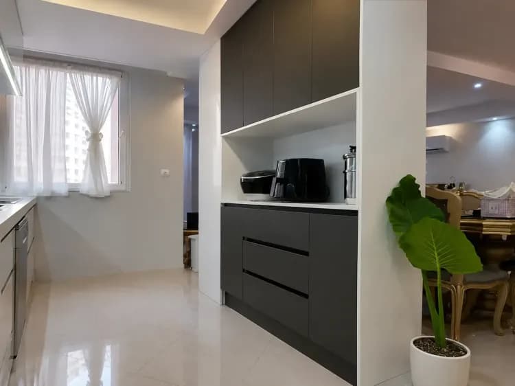 مایی از آشپزخانه مدرن با کابینت‌های سفید و طوسی، فضای مخصوص لوازم برقی و یک گلدان سبز در کنار ورودی سالن غذاخوری