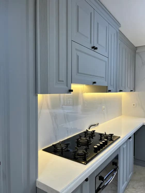 جزئیات کابینت نئوکلاسیک طوسی با ستون‌های شیاردار، گاز صفحه‌ای شیشه‌ای مشکی و بین‌کابینتی براق طرح ماربل با نور مخفی زرد.