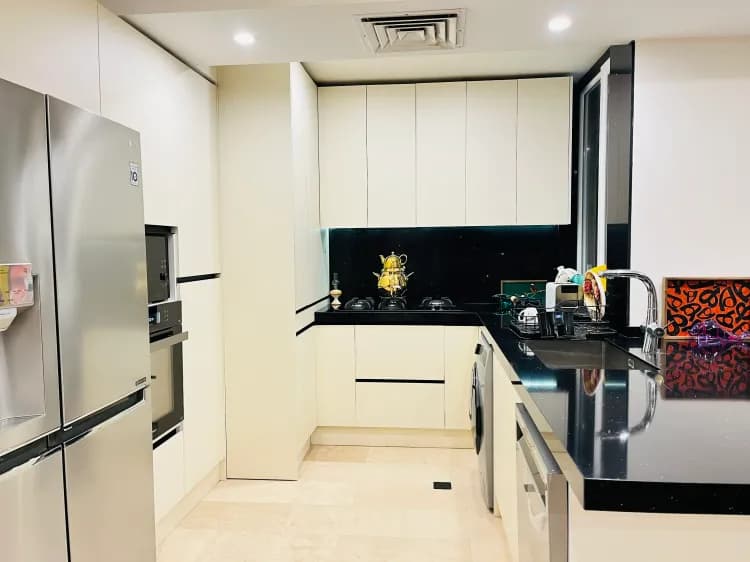 آشپزخانه مدرن U شکل با کابینتهای کرم روشن، صفحه و بینکابینتی مشکی، یخچال و لوازم استیل